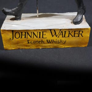 May include: A sculpture with the Johnnie Walker logo and the words "Scotch Whisky" on a gold-coloured base. The sculpture depicts a figure in black boots and a walking stick.
