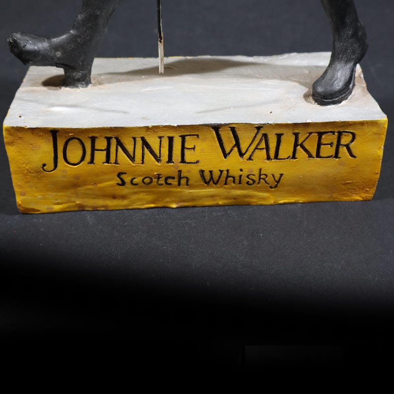 May include: A sculpture with the Johnnie Walker logo and the words "Scotch Whisky" on a gold-coloured base. The sculpture depicts a figure in black boots and a walking stick.