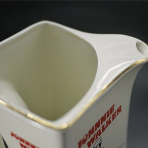 May include: Cream-colored ceramic pitcher with a square body and a curved handle. The pitcher has a gold trim along the rim and handle. The words "JOHNNIE WALKER" are printed in red on the side of the pitcher, along with a logo.