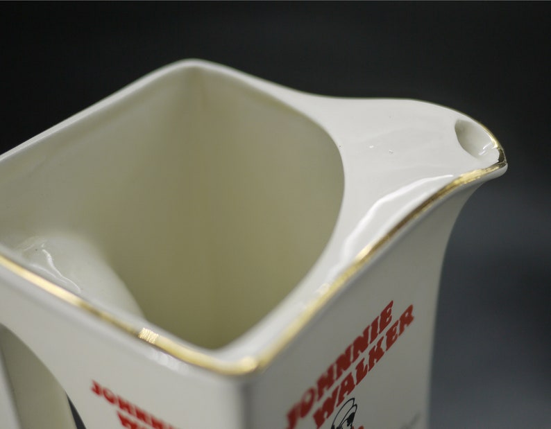 May include: Cream-colored ceramic pitcher with a square body and a curved handle. The pitcher has a gold trim along the rim and handle. The words "JOHNNIE WALKER" are printed in red on the side of the pitcher, along with a logo.