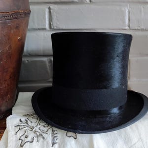 May include: A black top hat with a tall, cylindrical crown and a wide, curved brim. The hat features a smooth, dark finish and a matching black band encircling the base of the crown, providing a structured and formal appearance for this headwear.