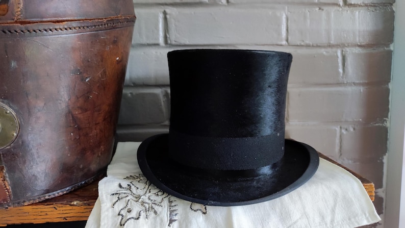 May include: A black top hat with a tall, cylindrical crown and a wide, curved brim. The hat features a smooth, dark finish and a matching black band encircling the base of the crown, providing a structured and formal appearance for this headwear.