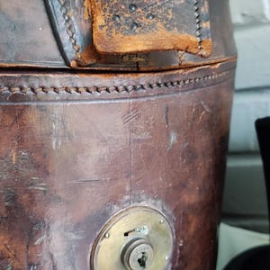 May include: A cylindrical brown leather hat box featuring visible stitching along the top edge, a worn leather tab closure, and a circular brass lock mechanism centered on the front. The leather surface shows signs of age, wear, and scuffs.