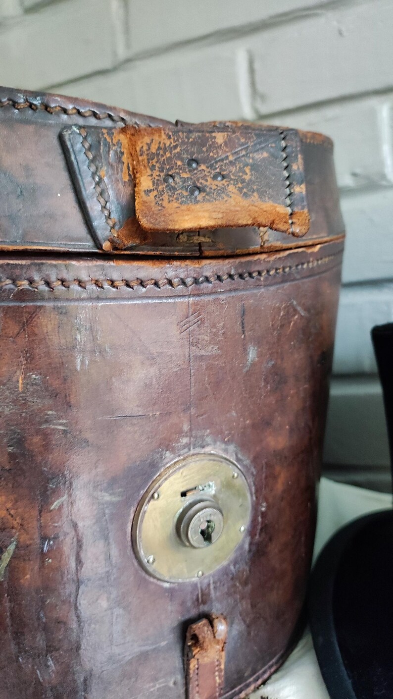 May include: A cylindrical brown leather hat box featuring visible stitching along the top edge, a worn leather tab closure, and a circular brass lock mechanism centered on the front. The leather surface shows signs of age, wear, and scuffs.