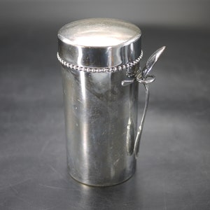 Peut inclure: Un récipient cylindrique argenté avec un couvercle et une petite poignée. Le récipient a un motif de perles autour du haut et un petit ustensile en forme de cuillère attaché sur le côté. L'article est en métal et a une finition brillante.