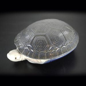 Peut inclure: Figurine de tortue en céramique avec une carapace bleu foncé et grise, ornée d'un motif hexagonal détaillé. La tête et les pieds de la tortue sont de couleur blanc crème, sur fond noir.