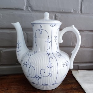 Peut inclure: Théière en céramique blanche avec couvercle et anse incurvée. La théière présente des détails côtelés verticaux et est ornée de motifs floraux et de vigne bleus. Le bec est long et fin. La théière est posée sur une surface en bois.