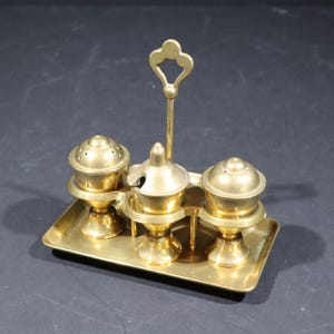 Puede incluir: Un juego de condimentos de latón antiguo que incluye un salero, un pimentero y un recipiente con tapa, todo sobre una bandeja rectangular. Un asa decorativa se eleva desde la bandeja. El juego tiene un acabado dorado pulido.
