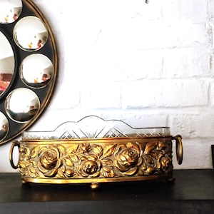 May include: An oval centerpiece bowl featuring a gold-toned metal frame with a detailed relief of roses and leaves. The bowl includes two side ring handles and a clear glass insert with a cut-glass diamond pattern along the upper rim.