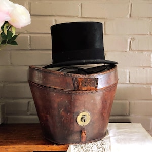 May include: A black top hat resting on top of a weathered, oval-shaped brown leather hat box. The leather container features visible stitching along the edges, a central brass locking mechanism, and a small leather tab closure on the front exterior.