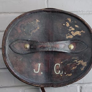 May include: A dark brown, oval-shaped leather hat box featuring a central handle secured with brass rivets. The surface shows signs of wear with distressed patches and the initials J. C. embossed in white lettering on the front face of the lid.