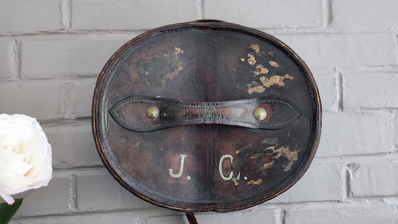 May include: A dark brown, oval-shaped leather hat box featuring a central handle secured with brass rivets. The surface shows signs of wear with distressed patches and the initials J. C. embossed in white lettering on the front face of the lid.