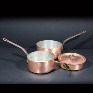 Pode incluir: Três panelas de cobre com cabos longos e uma tampa. As panelas têm um exterior de cobre brilhante e um interior prateado. A tampa tem uma pequena alça. O conjunto é exibido em uma superfície escura.