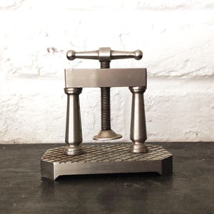 May include: A silver-toned metal book press featuring a central threaded screw mechanism with a horizontal handle, supported by two vertical pillars on a rectangular base with a textured, diamond-patterned surface for securing items.