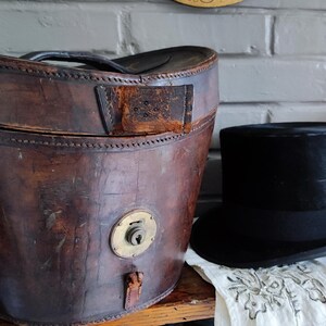 May include: A weathered brown leather hat box with a brass locking mechanism and a black handle, positioned next to a black felt top hat with a matching black ribbon band. The hat box features visible stitching along the lid and base edges.