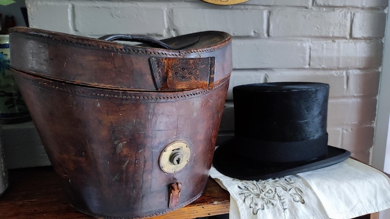 May include: A weathered brown leather hat box with a brass locking mechanism and a black handle, positioned next to a black felt top hat with a matching black ribbon band. The hat box features visible stitching along the lid and base edges.