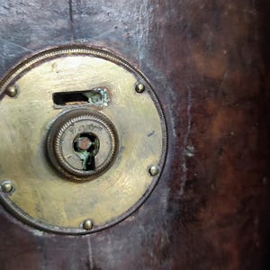 May include: A dark brown leather surface featuring a circular brass lock mechanism with a central keyhole and a rectangular slot above it. The brass plate is secured with small rivets and shows signs of oxidation around the central keyhole.