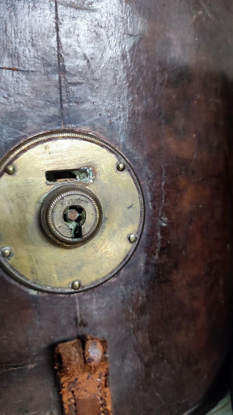 May include: A dark brown leather surface featuring a circular brass lock mechanism with a central keyhole and a rectangular slot above it. The brass plate is secured with small rivets and shows signs of oxidation around the central keyhole.