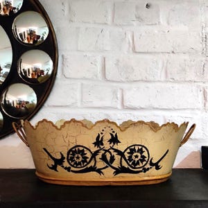 May include: An oval metal basin with a cream-colored crackle finish and a scalloped top edge. The exterior features a black painted floral and scrollwork design. Two small metal handles are attached to the sides of this decorative container.