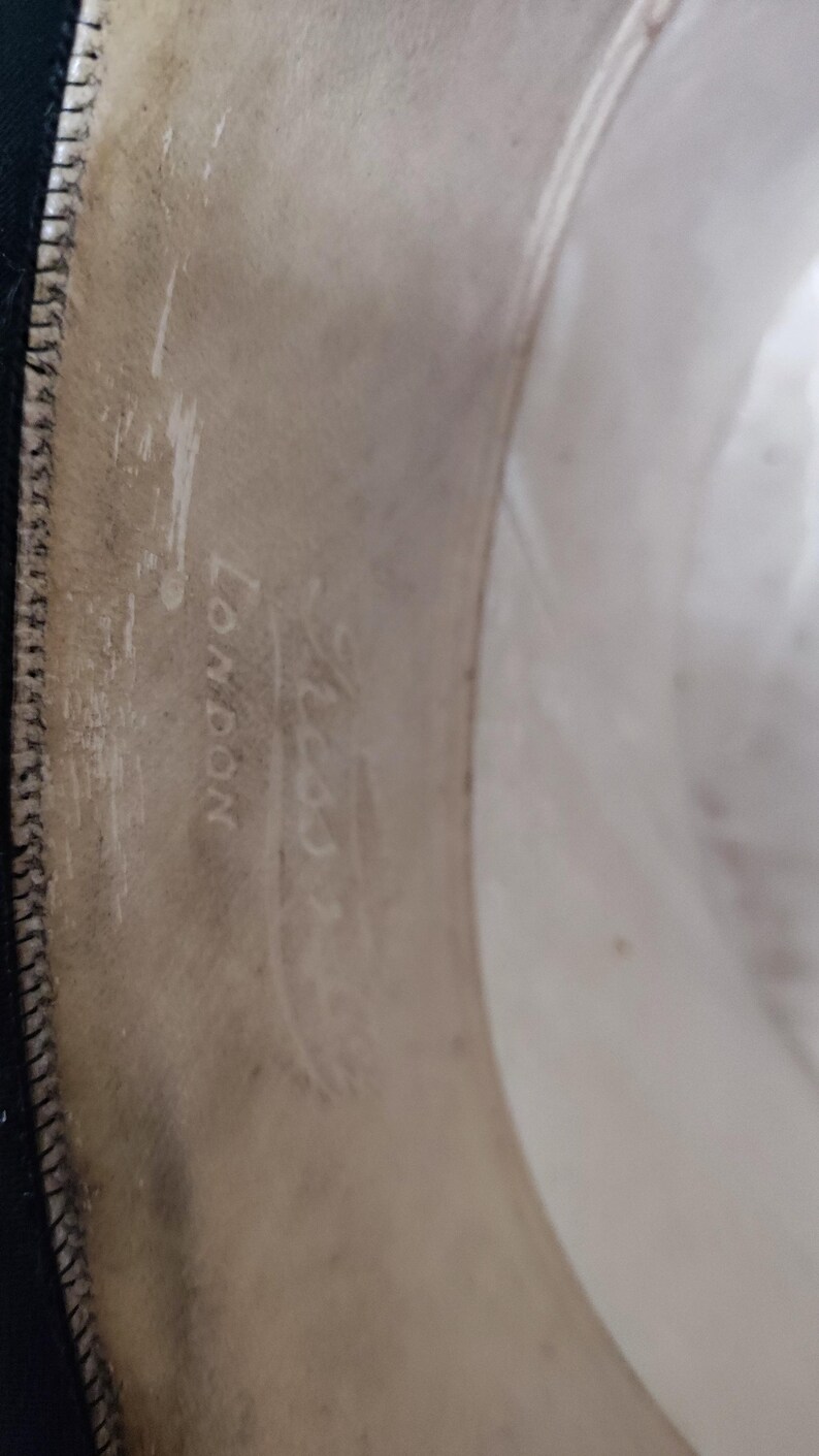 May include: The interior sweatband of a hat featuring a light beige leather lining with visible stitching along the edge. The text "LONDON" is clearly embossed on the material, accompanied by a faint, partially obscured brand name above it.