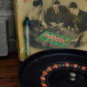 May include: A vintage roulette game set featuring a black plastic roulette wheel with numbered slots and a yellow cardboard box lid displaying a black and white illustration of people playing at a table with the text ROULETTE and a small green tube.