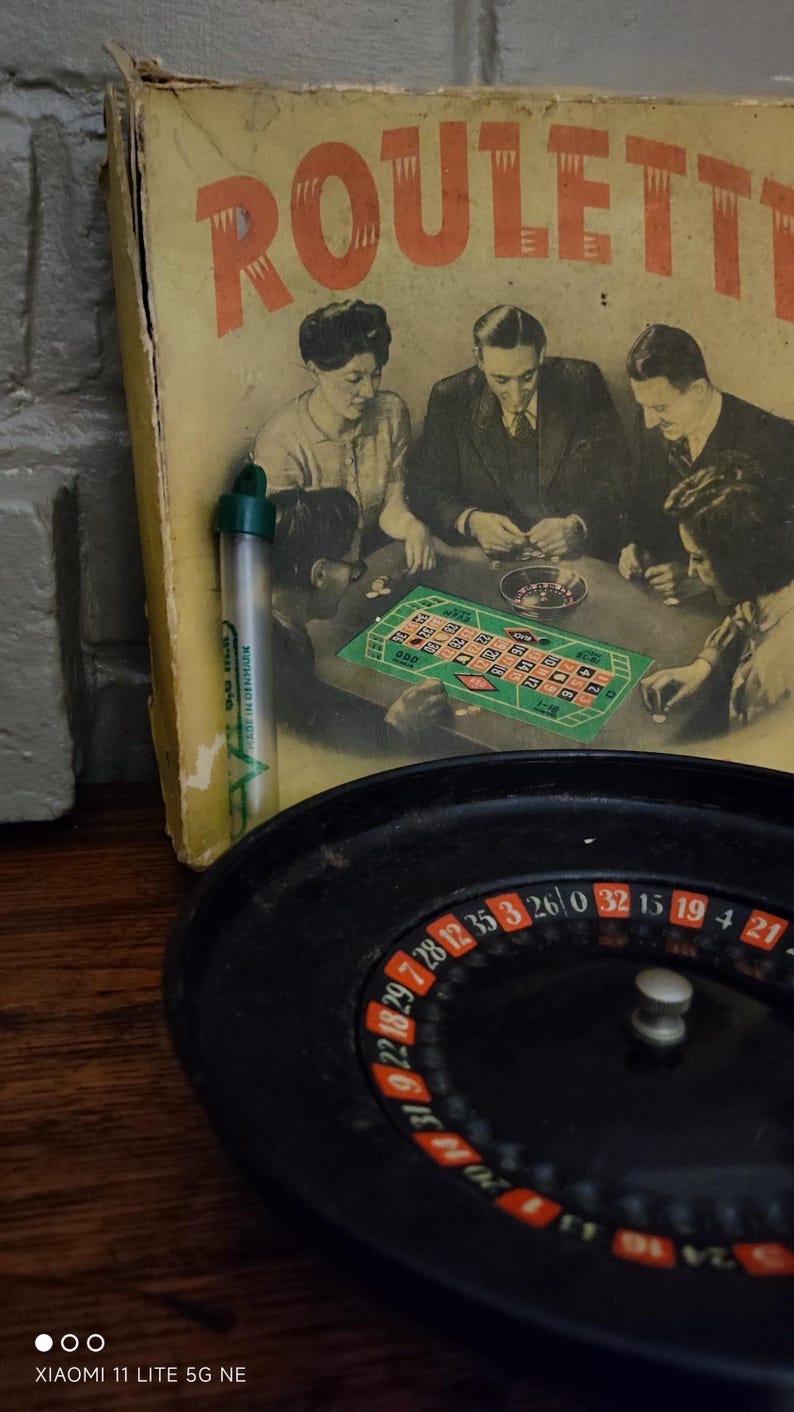 May include: A vintage roulette game set featuring a black plastic roulette wheel with numbered slots and a yellow cardboard box lid displaying a black and white illustration of people playing at a table with the text ROULETTE and a small green tube.