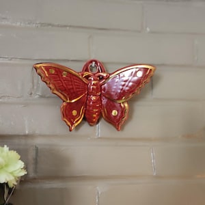 Puede incluir: Adorno de pared de cerámica roja con forma de mariposa y detalles dorados. La mariposa presenta intrincados diseños en las alas y pequeños puntos dorados. Está montada en una pared de ladrillo gris claro.