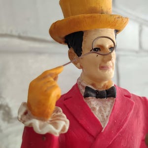 May include: A decorative figurine of a man in a red suit, yellow top hat, and orange gloves, holding a monocle. The figure has a black bow tie. The background is a white brick wall. A detailed ornament.