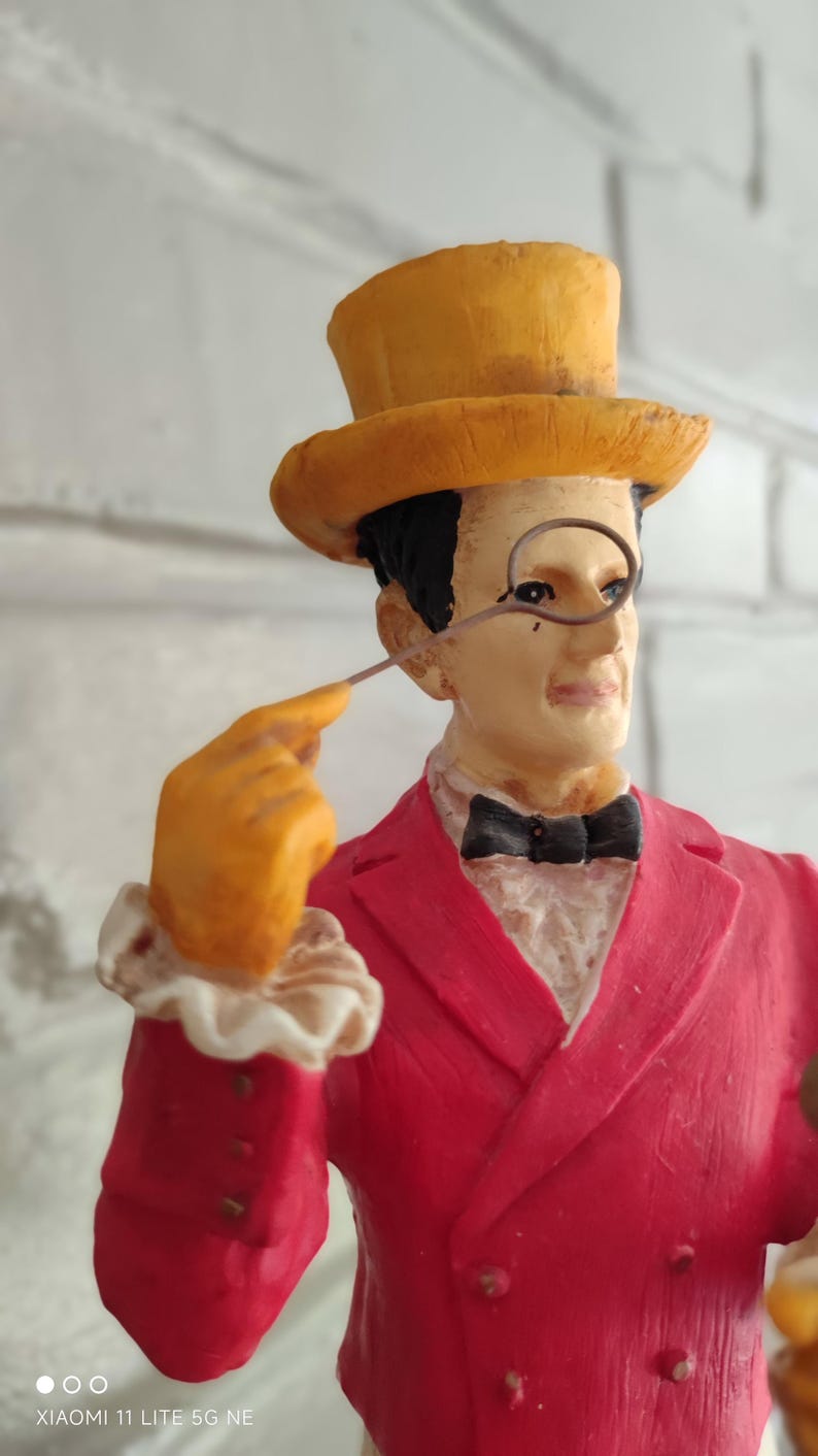 May include: A decorative figurine of a man in a red suit, yellow top hat, and orange gloves, holding a monocle. The figure has a black bow tie. The background is a white brick wall. A detailed ornament.