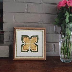 Puede incluir: Obra de arte enmarcada con un motivo floral. La imagen muestra una flor de cuatro pétalos en verde y dorado sobre un fondo blanco, dentro de un marco de madera. Un jarrón con rosas rosas es visible en el fondo. La obra está sobre una superficie de madera.