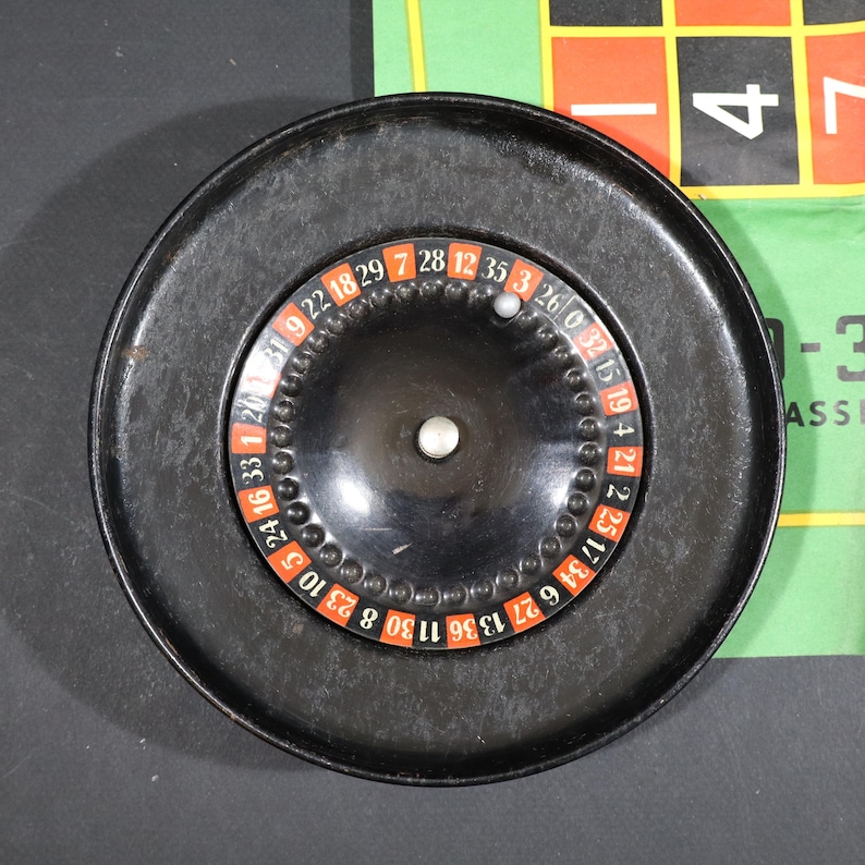 May include: A circular black roulette wheel featuring a central silver spindle and a small silver ball resting on the numbered track. The wheel displays alternating red and black numbered segments from 0 to 36, set within a wide black rim.