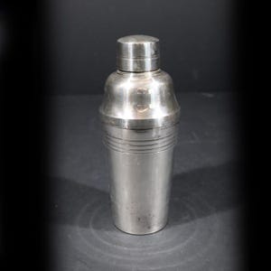 Peut inclure: Un shaker à cocktail en métal argenté composé de trois parties, avec un corps cylindrique, une section centrale effilée et un petit bouchon. La surface présente une finition lisse et polie avec des détails en anneaux horizontaux.