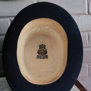 May include: A black top hat with a wide brim and a cream-colored interior lining. The inside features a central crest logo with the text "RESS & CO LONDON" printed in black ink. A small, light-colored ribbon bow is attached to the inner crown.