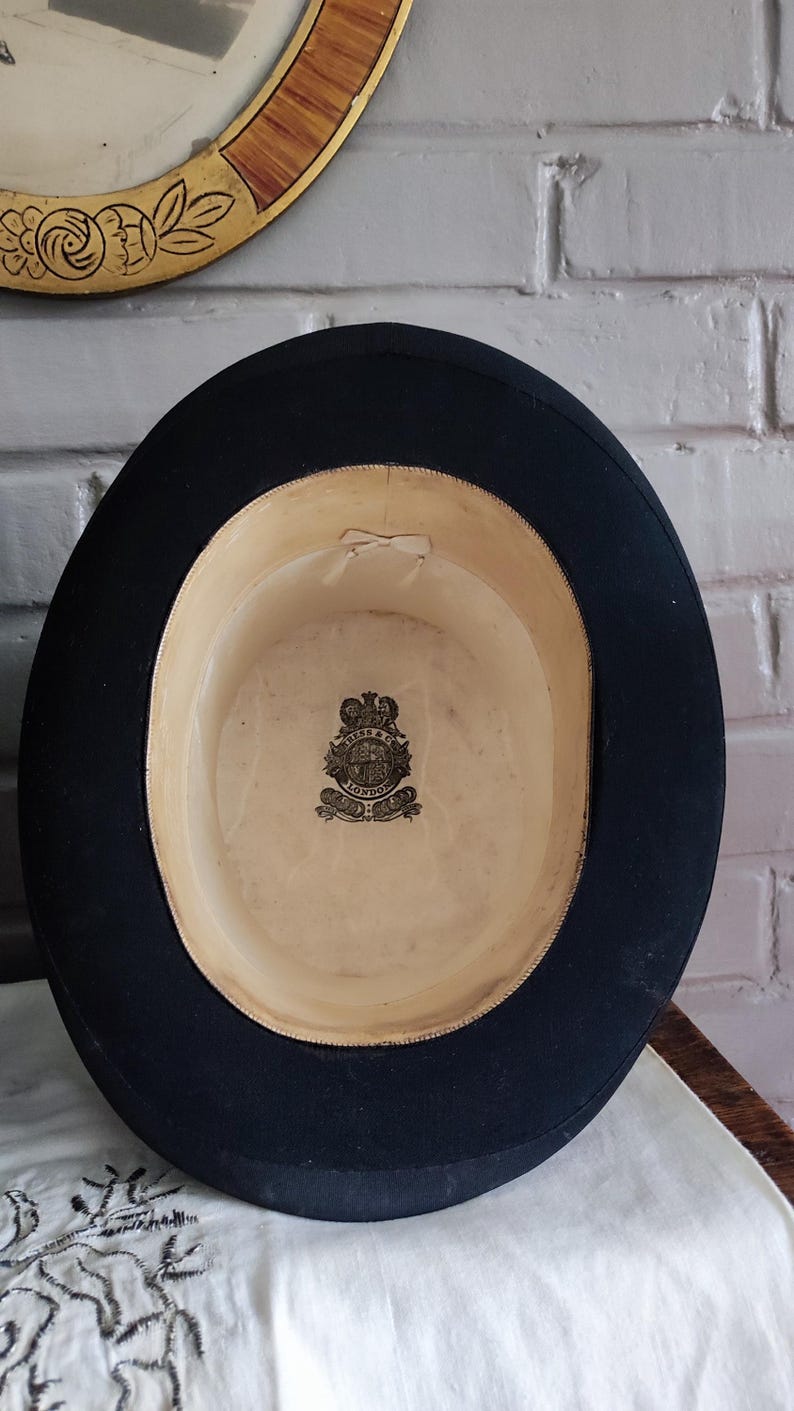 May include: A black top hat with a wide brim and a cream-colored interior lining. The inside features a central crest logo with the text "RESS & CO LONDON" printed in black ink. A small, light-colored ribbon bow is attached to the inner crown.