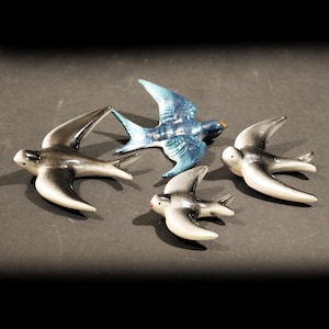 May include: A set of four ceramic bird wall ornaments in flight. Three birds feature a gradient of black to white, while one central bird is finished in a glossy blue glaze. Each bird is depicted with wings spread wide in a soaring position.