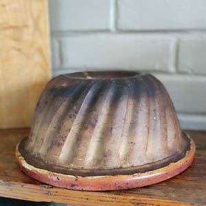 Peut inclure: Un moule à gâteau bundt rond et cannelé en céramique avec une finition tachetée brun foncé et beige vieillie. Le moule présente un large bord plat à la base et un centre creux, offrant un aspect rustique avec une texture de surface visible.