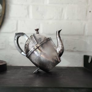 Peut inclure: Une théière en argent vintage avec un bec verseur, une anse et un couvercle incurvés. La théière repose sur un petit support, et la surface a une finition mate foncée. L'arrière-plan présente un mur de briques blanches.