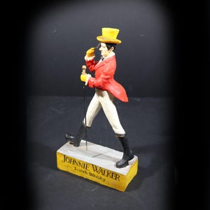 May include: Figurine of a man in a red coat, yellow top hat, and white trousers, holding a cane. The figurine is walking on a yellow base with the words "JOHNNIE WALKER Scotch Whisky".