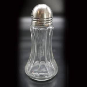 Peut inclure: Un saupoudreur à sel en verre transparent avec un dessus argenté. Le saupoudreur a un design cannelé et une base ronde.