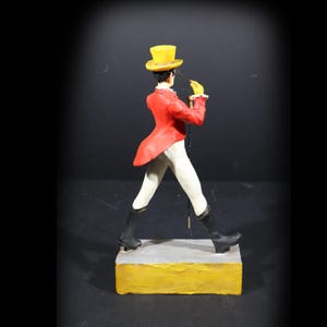 May include: A figurine of a person in a red jacket, white trousers, and black boots, holding a riding crop. The figure wears a yellow top hat and gloves. The figurine stands on a yellow and grey rectangular base.