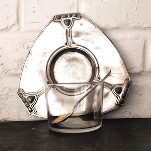 May include: A silver-toned triangular metal saucer with geometric detailing and dark inlays, paired with a clear glass tumbler and a small metal spoon. The saucer features a central circular indentation and decorative accents at each corner.