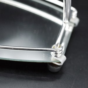 May include: Close-up of a chrome-plated, two-tiered serving tray with a mirrored surface. The tray has a rounded shape with a curved handle and small, rounded feet. The design is simple and elegant.