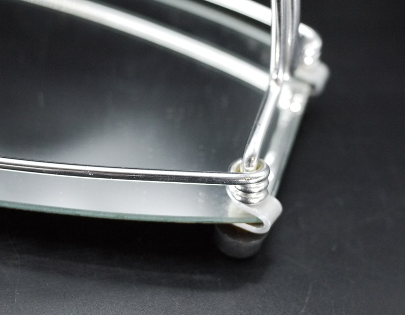 May include: Close-up of a chrome-plated, two-tiered serving tray with a mirrored surface. The tray has a rounded shape with a curved handle and small, rounded feet. The design is simple and elegant.