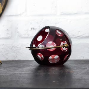 Peut inclure: Un cendrier en verre rouge rubis avec des pois blancs et un bord en laiton. Le cendrier est rond avec un dessus légèrement aplati et repose sur une surface sombre. Un objet décoratif doré est en arrière-plan.