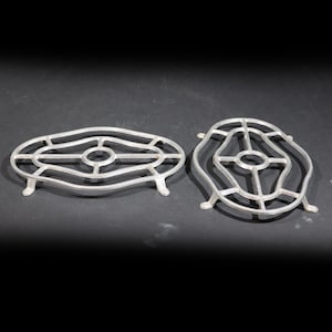 Puede incluir: Dos salvamanteles de metal plateado. Uno es de forma ovalada con un círculo central y un diseño calado decorativo. El otro tiene forma redondeada con un diseño calado similar. Ambos tienen pequeños pies.