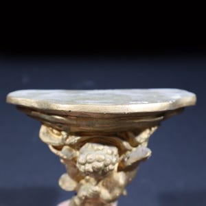 May include: A gold-colored decorative shelf with an ornate design. The shelf is shaped like a half-circle and has a raised edge. The shelf is supported by a sculpted figure.