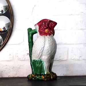 Peut inclure: Pichet en céramique en forme de perroquet, avec un corps blanc et rouge et des accents verts. La poignée verte imite une tige de bambou. La base est un mélange de vert, de jaune et de marron.