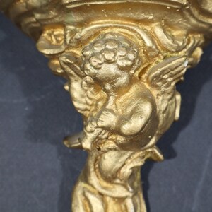 May include: A gold-colored metal wall sconce with a cherub figure holding a flower. The sconce has a decorative, ornate design.