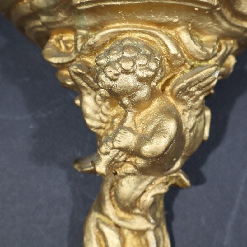 May include: A gold-colored metal wall sconce with a cherub figure holding a flower. The sconce has a decorative, ornate design.