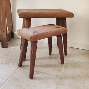 Peut inclure: Deux tabourets en bois brun avec des plateaux rectangulaires et quatre pieds fuselés. Les tabourets présentent une finition en grain de bois naturel avec des nœuds circulaires visibles sur les assises. Le design est simple, avec un grand tabouret derrière.